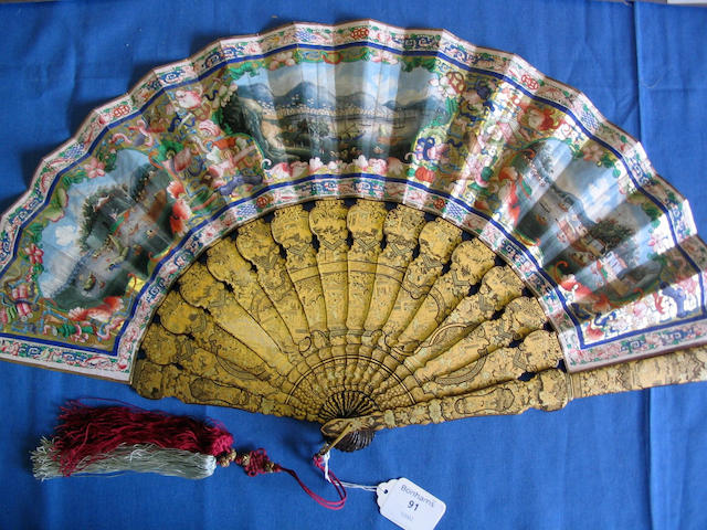 A good early 19th Century Chinese export fan The bright gold and black lacquer guards and sticks well-decorated with figures on terraces amid panels of birds, animals and objects, the leaf painted to one side with numerous ivory faced figures arranged on terraces and within defined areas, two horsemen to the fore, within a trellis, objects, animal and floral border, the reverse of the leaf painted with three cartouche panels, two of river scenes, the other a trade port, within brightly coloured objects, leaf scroll flowers and within a formal bird and trellis border.