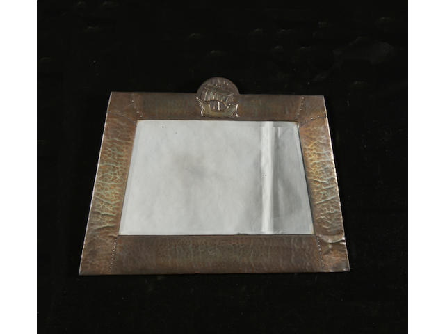 An Arts and Crafts beaten metal framed bevelled wall mirror, with galleon crest and tapered sides, 90cm wide x 64cm high.